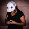 Person wearing a white cap with a logo against a brick wall.